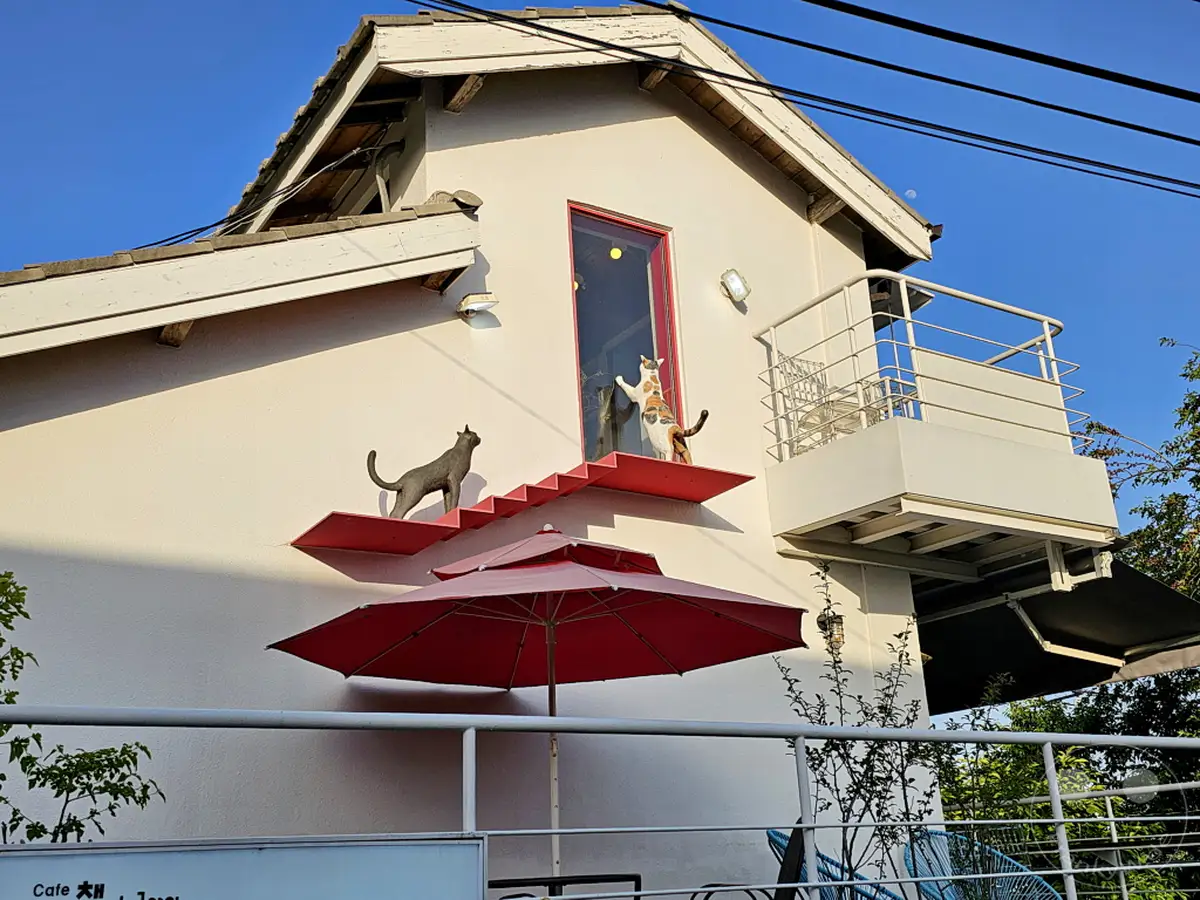 Eine schlichte weiße Hausfassade in Seoul, Südkorea, wird durch skurrile Katzenfiguren auf roten Regalen belebt, die wie an einer Treppe auf und ab klettern. Ein Sonnenschirm wirft einen Schatten und eine kleine Terrasse bietet einen Blick auf die Umgebung, während der blaue Himmel und die klare Sonne eine helle und einladende Atmosphäre schaffen.