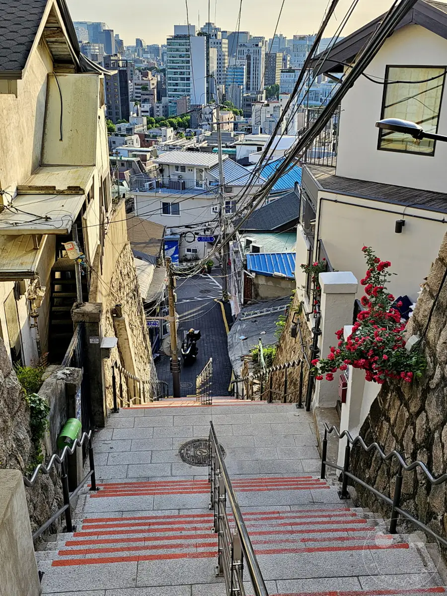 Eine steile Steintreppe mit roten Markierungen führt hinunter in eine enge Gasse im Ihwa Mural Village in Seoul, Südkorea. Von oben bietet sich ein Blick über die Dächer der Stadt und aufgestellte Stromleitungen, die das tägliche Leben widerspiegeln. Die Szenerie strahlt eine authentische und charmante Atmosphäre eines lebendigen Stadtviertels aus.