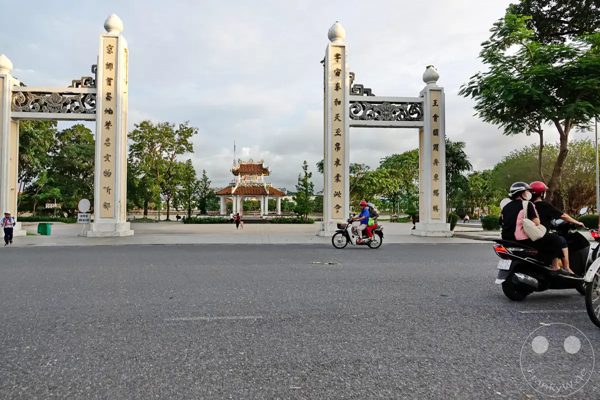 Vor einem imposanten Torbogen im Thuong Bac Park in Hue, Vietnam, fährt ein Motorroller mit zwei Insassen vorbei. Im Hintergrund ist ein traditionelles Gebäude mit mehreren Dächern zu sehen, das von Bäumen und einer ruhigen Atmosphäre umgeben ist. Die Szene vermittelt einen Eindruck von Alltag und kultureller Präsenz in dieser historischen vietnamesischen Stadt.