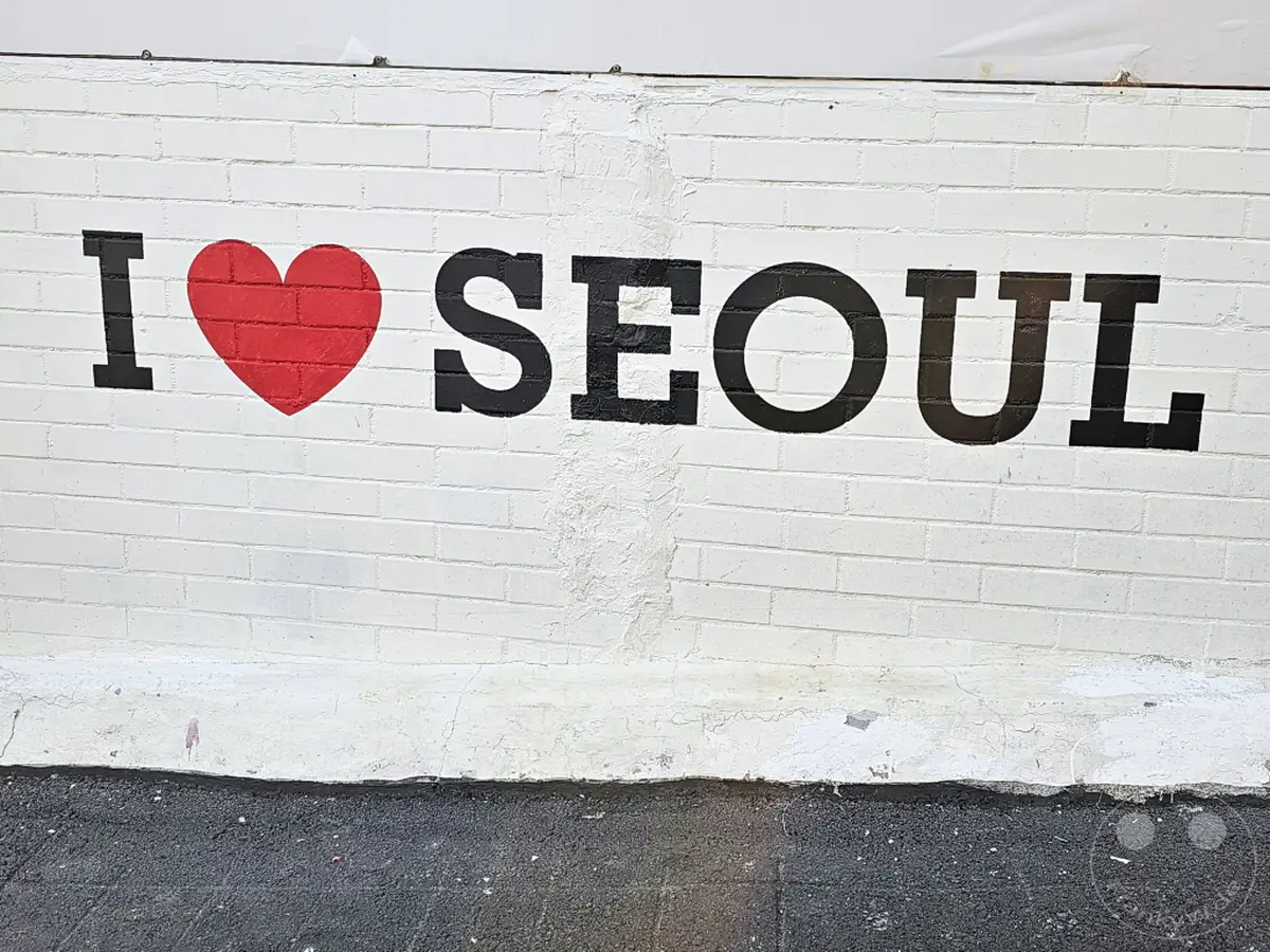 Ein rot gemaltes Herz und schwarze Großbuchstaben formen die Aussage "I LOVE SEOUL" auf einer weißen Ziegelmauer in Südkorea. Das Kunstwerk befindet sich in der Nähe des Ihwa Mural Village und strahlt eine positive und einladende Atmosphäre aus. Die raue Textur der Wand und der darunter liegende Asphaltboden verleihen der Szene einen authentischen urbanen Charakter.