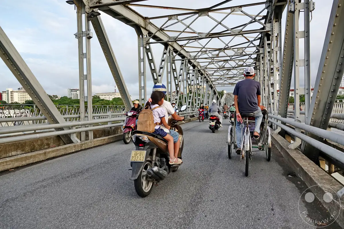 Eine belebte Szene auf der historischen Truong Tien Brücke in Hue, Vietnam, wo Motorroller und eine Fahrradrikscha den Verkehr anführen. Der morgendliche oder abendliche Himmel ist von leichten Wolken bedeckt, während die Menschen ihre täglichen Wege auf dieser ikonischen Flussüberquerung zurücklegen. Die Szenerie strahlt ein Gefühl von Alltagsleben und lokaler Mobilität in einer Stadt voller Geschichte aus.