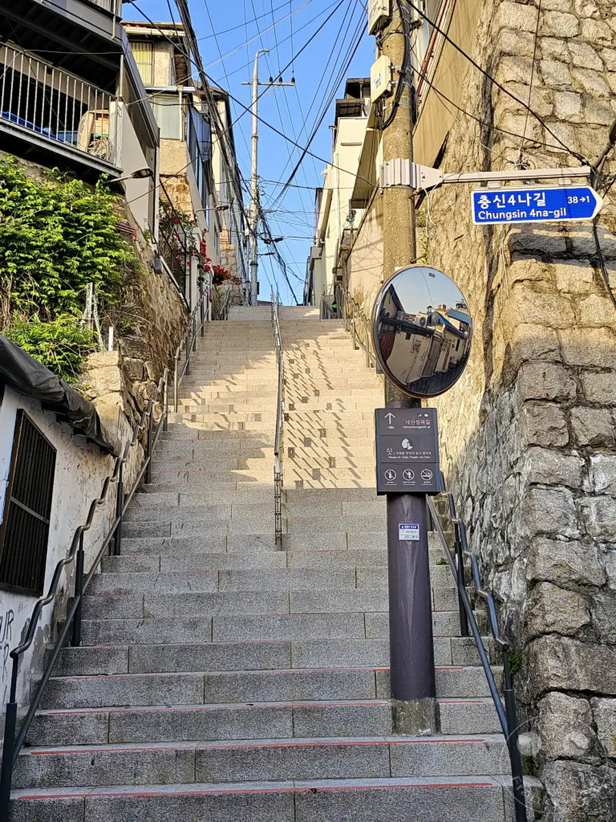Eine steile Steintreppe führt bergauf durch das Ihwa Mural Village in Seoul, Südkorea. An den Seiten sind traditionelle koreanische Häuser und steinerne Mauern zu sehen, die von einem Netz aus Stromleitungen überspannt werden. Die sonnige Atmosphäre und die Architektur verleihen der Szene einen besonderen Charme.