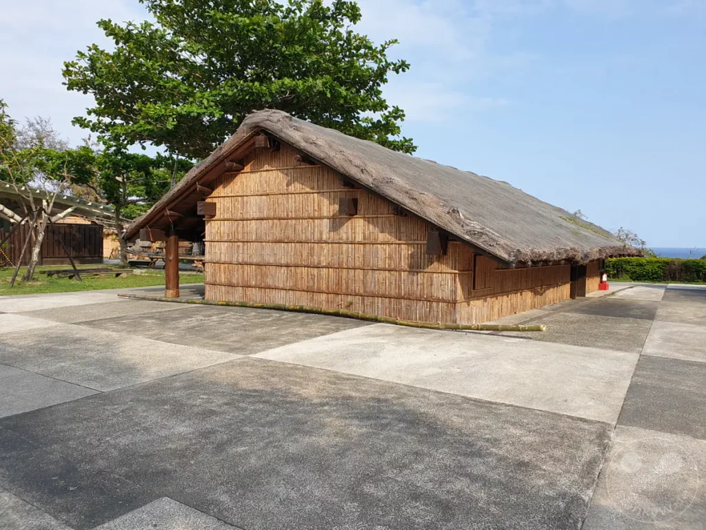 Eine traditionelle Bambushütte steht auf dem Gelände des Amis Folk Centers in Taiwan. Der Blick schweift über das Meer und eine üppig grüne Landschaft, die von einem strahlend blauen Himmel überdacht wird. Die Szene strahlt eine friedliche und entspannte Urlaubsatmosphäre aus.