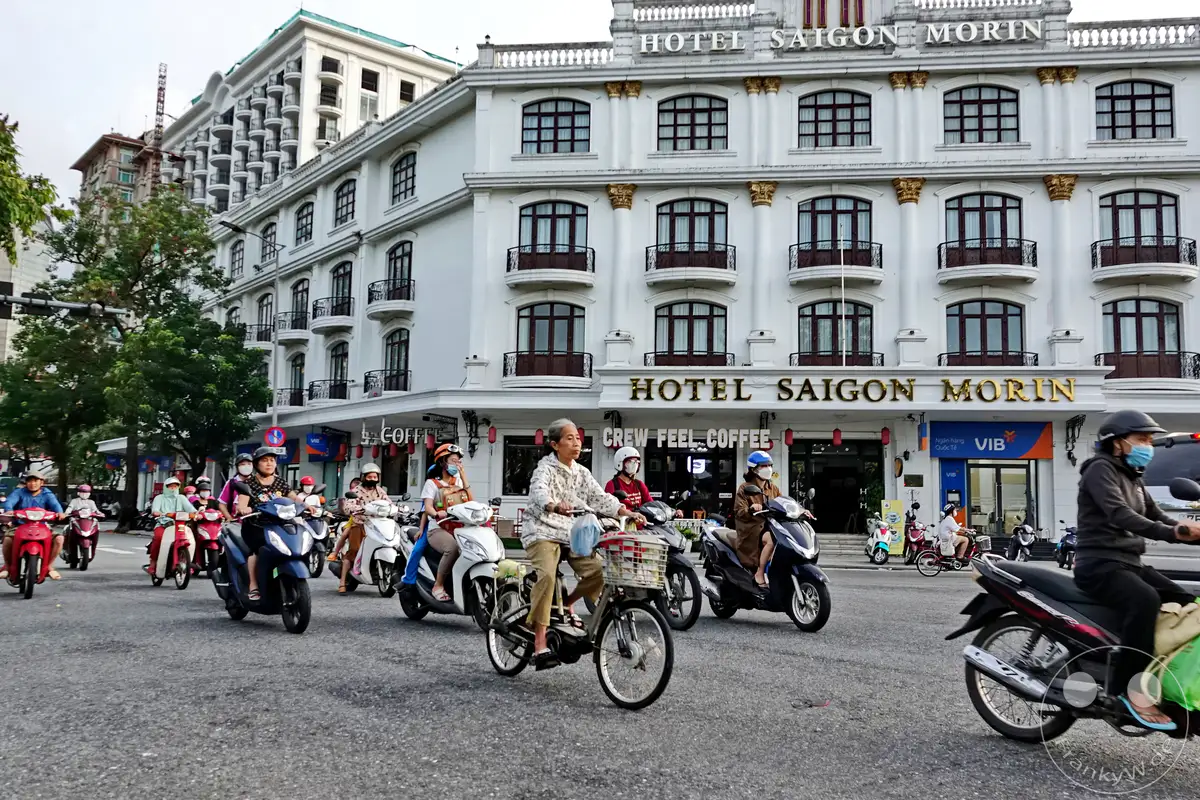 Motorrollerfahrer bewegen sich tagsüber durch eine belebte Straße in Hue, Vietnam, mit dem markanten Hotel Saigon Morin im Hintergrund. Die Architektur des Hotels, ein Überbleibsel einer vergangenen Ära, kontrastiert mit dem modernen Verkehrsgeschehen. Die Szene vermittelt einen Eindruck vom täglichen Leben und dem urbanen Charakter der Stadt.