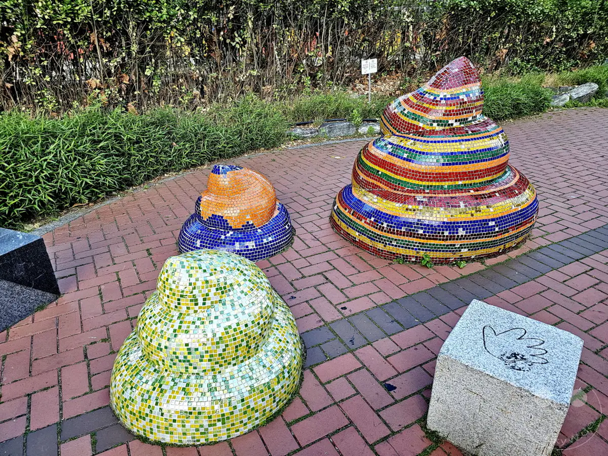 Im Daehangno district in Seoul, Südkorea, stehen bunte Mosaik-Skulpturen, die an Kot erinnern, auf einem gepflasterten Weg. Die lebendigen Farben und die humorvolle Formgebung schaffen eine verspielte und unerwartete Atmosphäre im öffentlichen Raum. Eine Gedenkstele mit einer Handabdruck-Gravur befindet sich ebenfalls in der Nähe, was auf eine künstlerische Gestaltung des Geländes hindeutet.