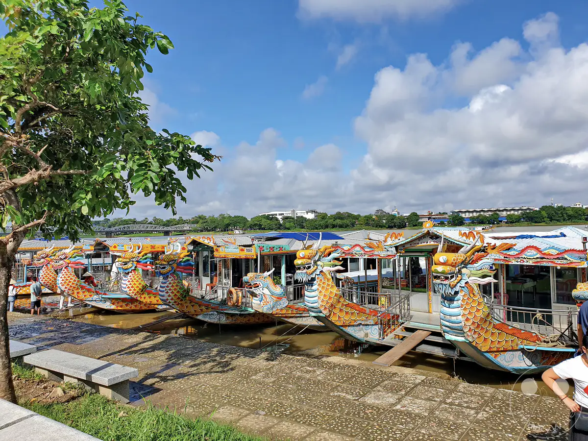 Mehrere traditionelle Drachenboote liegen im Sonnenschein am Ufer des Parfümflusses in Hue, Vietnam. Die farbenfrohen Boote mit Drachenköpfen und aufwändigen Verzierungen warten auf Touristen, um die Stadt und ihre Umgebung vom Wasser aus zu erkunden. Die Atmosphäre ist ruhig und einladend, perfekt für eine malerische Bootsfahrt.