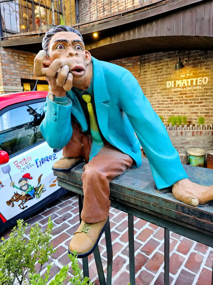 Eine lebensechte Skulptur eines Mannes mit einem neugierigen Ausdruck sitzt auf einer Bank vor einem Gebäude mit dem Namen "DI MATTEO Pizzeria & Trattoria". Das bunte Kunstwerk im Stadtteil Daehangno in Seoul, Südkorea, zieht die Aufmerksamkeit auf sich und verleiht der Straße einen künstlerischen Akzent. Die Szene vermittelt eine verspielte und einladende Atmosphäre, die zum Verweilen einlädt.