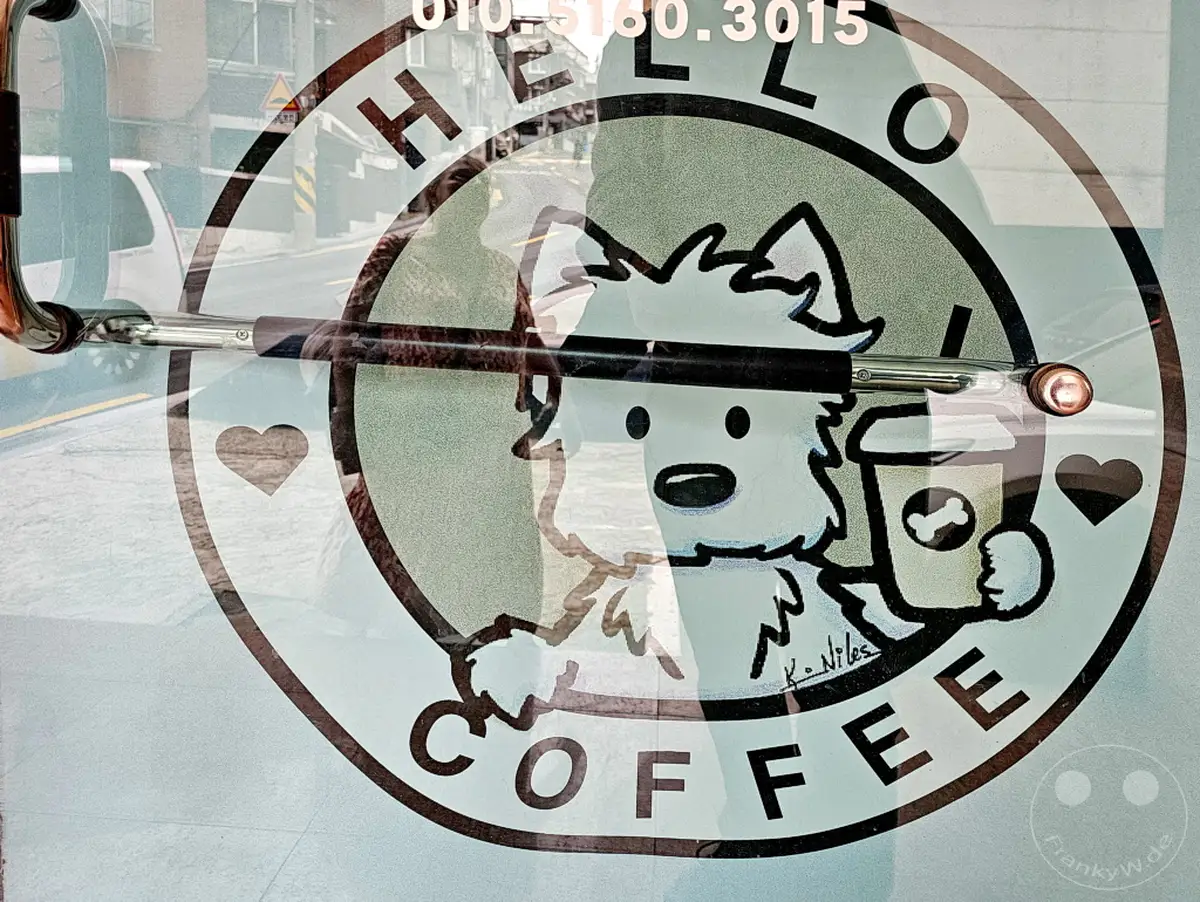 Eine belebte Wäscherei mit integriertem Café in Seoul, Südkorea, wirbt mit einem charmanten Hundelogo für ihr 'Hello Coffee'. Das Logo auf der Glastür zeigt einen niedlichen Hund, der einen Kaffeebecher hält, und lädt zu einer gemütlichen Pause ein. Die Umgebung deutet auf ein urbanes Umfeld hin, in dem Dienstleistung und Genuss verschmelzen.