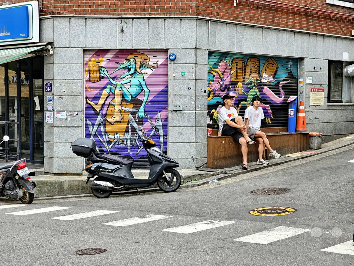 Zwei junge Männer sitzen entspannt auf einer Holzbank vor einer Hausfassade mit auffälligen Graffitis in Südkorea. Eine schwarze Vespa steht geparkt auf der Straße vor dem Gebäude, während im Hintergrund eine weitere Roller zu sehen ist. Die Szene vermittelt eine alltägliche urbane Atmosphäre mit künstlerischen Akzenten in Seoul.