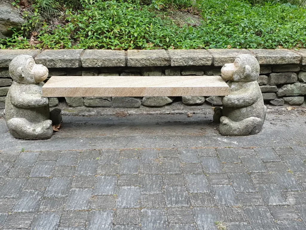 Zwei steinerne Affenfiguren stützen eine schlichte Steinbank inmitten einer Gartenlandschaft in Taiwan. Die ruhige Szenerie wird durch die angrenzende Steinmauer und üppige Vegetation unterstrichen. Die Sitzgelegenheit lädt zum Verweilen in der naturverbundenen Atmosphäre ein.