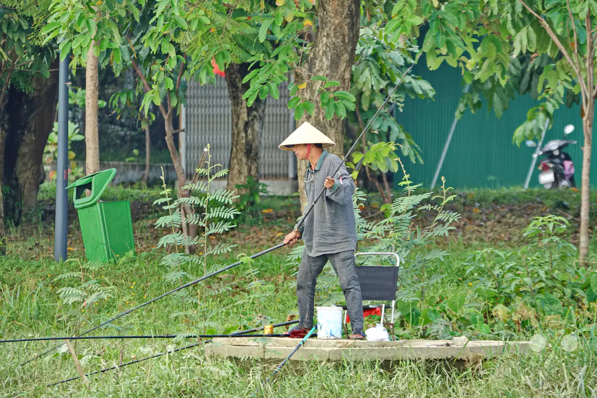 In Vietnam, nahe Hue, verbringt ein Angler seine Zeit am Flussufer und wirft seine Angel aus. Die ruhige Szene, geprägt von üppigem Grün und der Natur, strahlt Gelassenheit und Geduld aus. Ein Motorroller im Hintergrund und ein Abfalleimer am Wegesrand deuten auf eine urbane Nähe trotz der idyllischen Kulisse hin.