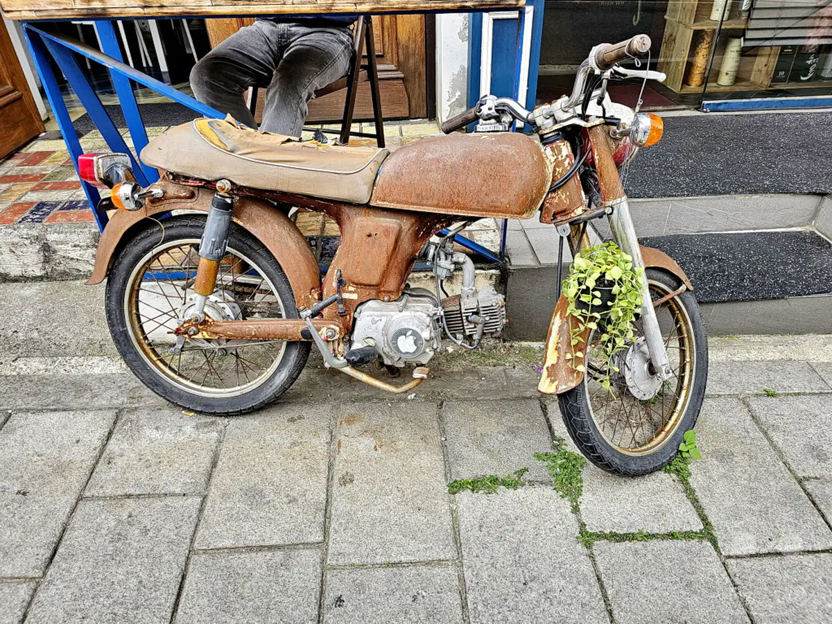 Ein stark verrostetes Moped steht auf einem gepflasterten Bürgersteig im Yongsan District von Seoul, Südkorea. Eine blaue Stahlkonstruktion stützt einen Tisch darüber, während eine hängende Pflanze am Vorderrad angebracht ist. Die Szene strahlt eine nostalgische und etwas melancholische Atmosphäre aus, die vom Verfall und der ungewöhnlichen Dekoration des Mopeds geprägt ist.