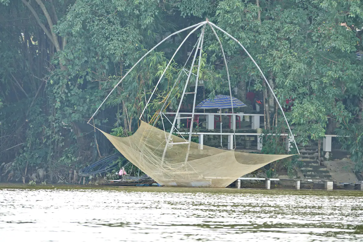 Ein großes, traditionelles Fischernetz steht am Ufer des Parfümflusses in Hue, Vietnam. Das Netz aus hellem Stoff ist auf einem Holzgerüst aufgespannt und ragt über das Wasser hinaus, während im Hintergrund dichter Baumwuchs und ein kleines Gebäude zu sehen sind. Die Szene strahlt eine ruhige Atmosphäre aus, die durch das trübe Wetter und die Spiegelungen auf der Wasseroberfläche verstärkt wird.