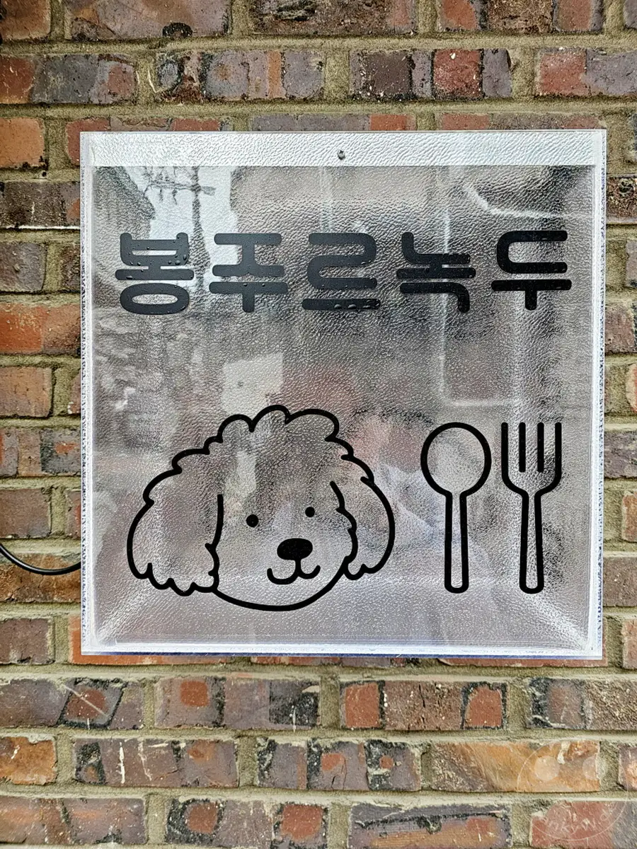 Ein Schild mit koreanischer Schrift, dem Wort 'Bongjurnukdu' und einem süßen Cartoon-Hund mit einer Gabel und einem Löffel ist an einer Ziegelwand befestigt. Das Schild scheint zu einem Lokal im Yongsan District von Seoul, Südkorea, zu gehören und vermittelt eine freundliche und einladende Atmosphäre. Die klare und texturierte Oberfläche des Schildes reflektiert die Umgebung und fügt dem Bild eine interessante visuelle Ebene hinzu.