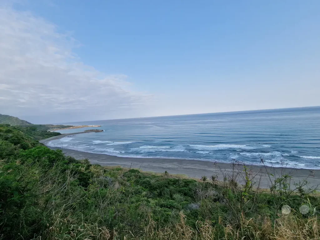 Eine weite Meeresansicht von einer grünen Küste in Taiwan. Sanfte Wellen brechen sich am dunklen Sandstrand, während die Sonne den Horizont in einem weiten Himmelsblau einfärbt. Üppige Vegetation bedeckt die Hügel und bietet einen atemberaubenden Blick auf die natürliche Schönheit der Küstenlandschaft.