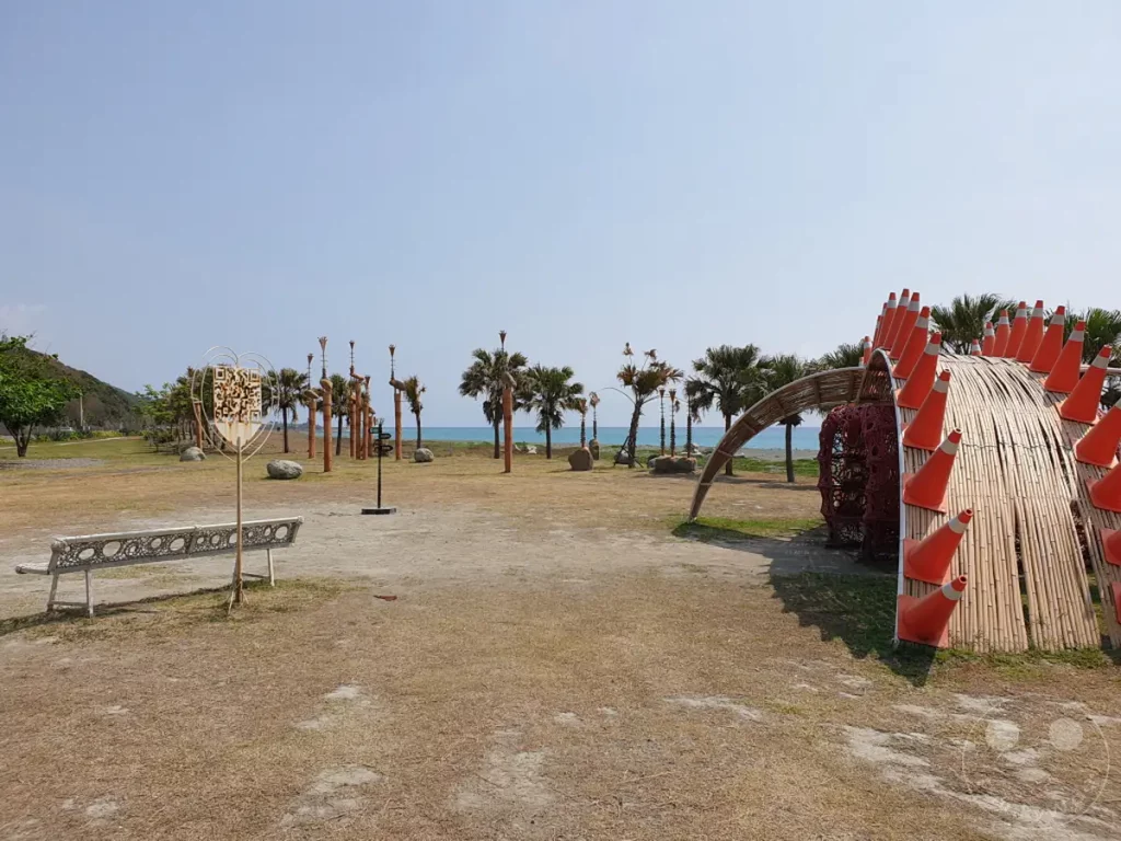 Die weite Küstenlandschaft von Taiwan im Dawu Coast Park bietet einen offenen Himmel über saftigen Palmen und dem azurblauen Meer. Eine interessante Kunstinstallation mit roten Kegeln und Bambusstrukturen dominiert den rechten Teil des Bildes, während im Vordergrund eine dekorative Bank zum Verweilen einlädt. Die ruhige Atmosphäre wird durch die klare Luft und das helle Sonnenlicht unterstrichen.