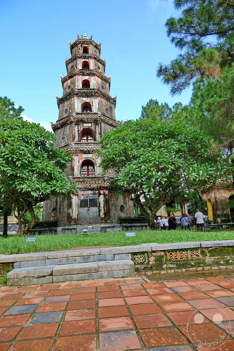 Die siebenstöckige Thien Mu Pagode, auch bekannt als Pagode der Himmlischen Dame, steht majestätisch vor einem klaren blauen Himmel in Hue, Vietnam. Üppige grüne Bäume säumen das Gelände, während Besucher die historische Stätte erkunden. Der Boden ist mit rötlichen Ziegelsteinen gepflastert, was dem gesamten Bild eine warme und authentische Atmosphäre verleiht.