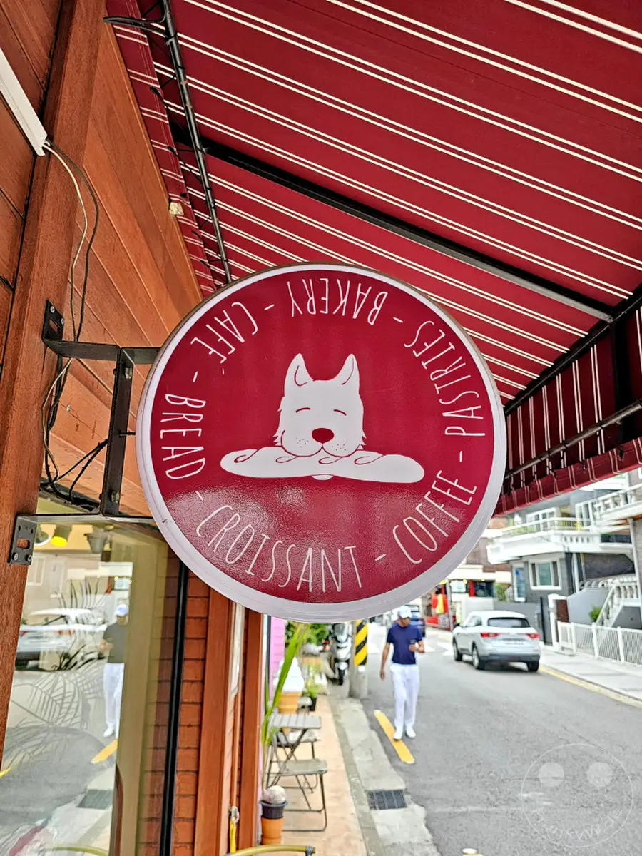 Ein rundes, rotes Schild mit einem stilisierten Hund und den Wörtern "Bakery", "Cafe", "Bread", "Pastries", "Croissant" und "Coffee" hängt vor einem Holzgebäude. Die rote Markise über dem Schild wirft Schatten und unterstreicht die charmante Atmosphäre des Ortes im Yongsan District von Seoul. Passanten und Autos sind auf der Straße im Hintergrund zu sehen, die ein lebhaftes Stadtbild von Südkorea widerspiegeln.