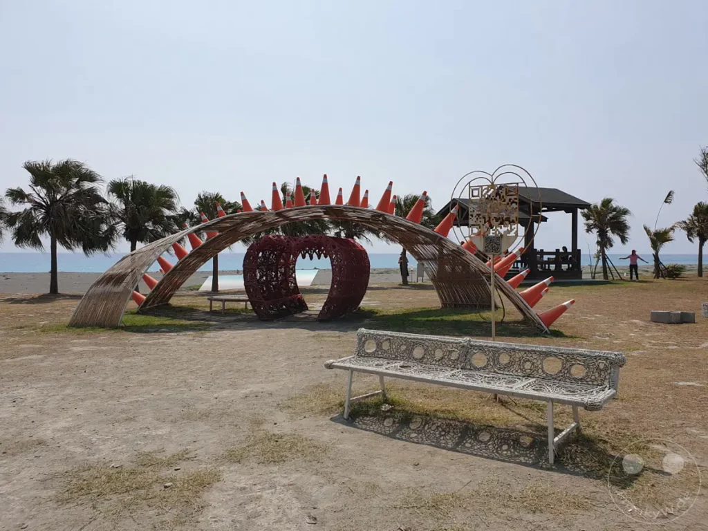 In Taiwan, am Dawu Coast Park, erstreckt sich eine bizarre Kunstinstallation am Strand, die an ein Monstermaul erinnert, flankiert von Palmen und dem weiten Meer. Eine weiße, ornamentale Bank lädt zum Verweilen ein, während im Hintergrund ein kleiner Pavillon Schatten spendet. Die Szene strahlt eine ruhige, sonnige Atmosphäre aus, die das Zusammenspiel von Natur und menschlicher Kreativität einfängt.