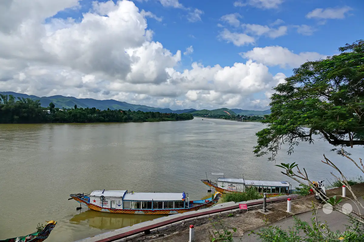 Ein breiter, schlammiger Fluss fließt durch eine grüne Landschaft mit sanften Bergen am Horizont. Mehrere traditionelle Drachenboote liegen am Ufer vor Anker, einige sind mit bunten Verzierungen geschmückt. Die bewölkte, aber helle Himmelskulisse verleiht der Szene eine friedliche und malerische Atmosphäre in Vietnam, Hue.