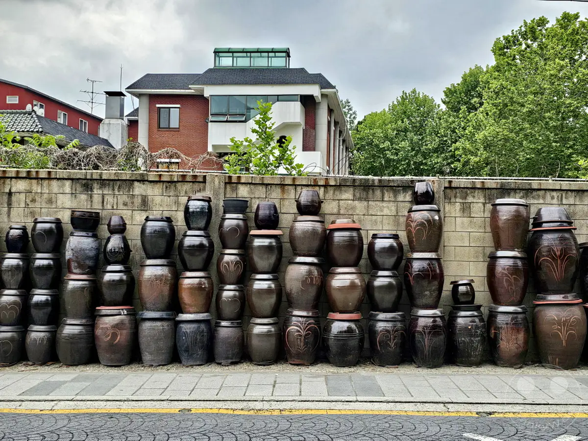 Eine Mauer aus Backsteinen in Südkorea, Seoul, im Yongsan District, ist gesäumt von einer Vielzahl traditioneller Keramikkrüge, die in kunstvollen Reihen gestapelt sind. Hinter der Mauer erheben sich ein Backsteingebäude mit moderner Architektur und üppig grüne Bäume, die eine atmosphärische Kulisse bilden. Die Anordnung der Krüge erzeugt ein Gefühl von Geschichte und Handwerkskunst in dieser städtischen Umgebung.