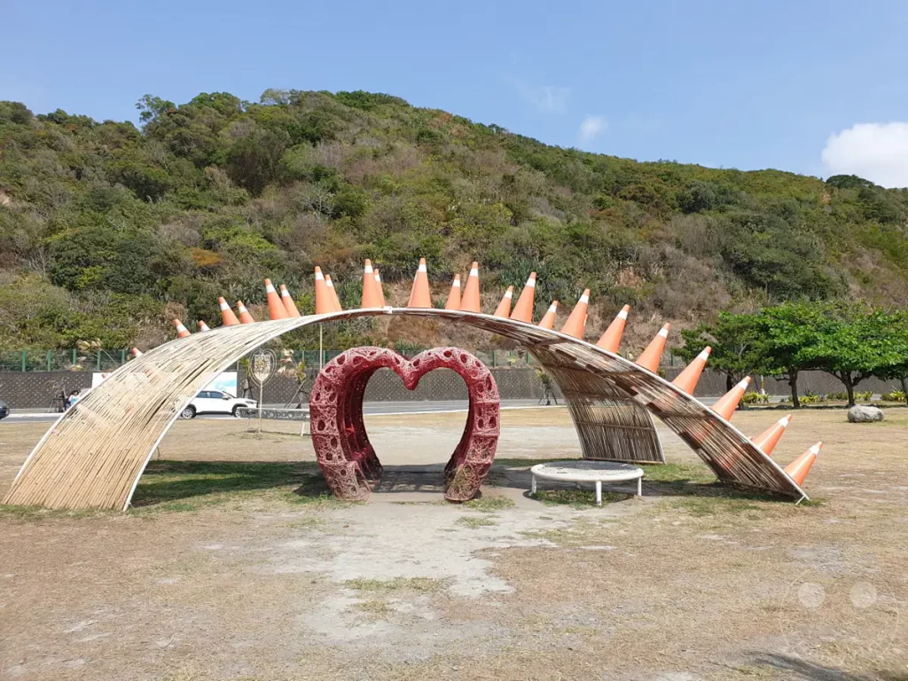 Im Dawu Coast Park in Taiwan steht eine künstlerische Herzskulptur, umgeben von einer beeindruckenden Naturkulisse mit grünen Hügeln und dem blauen Himmel. Die mit Verkehrskegeln verzierte Holzkonstruktion schafft eine surreale und einladende Atmosphäre an der Küste. Der sonnige Tag und die weitläufige Landschaft laden zu einem entspannten Besuch und zum Verweilen ein.