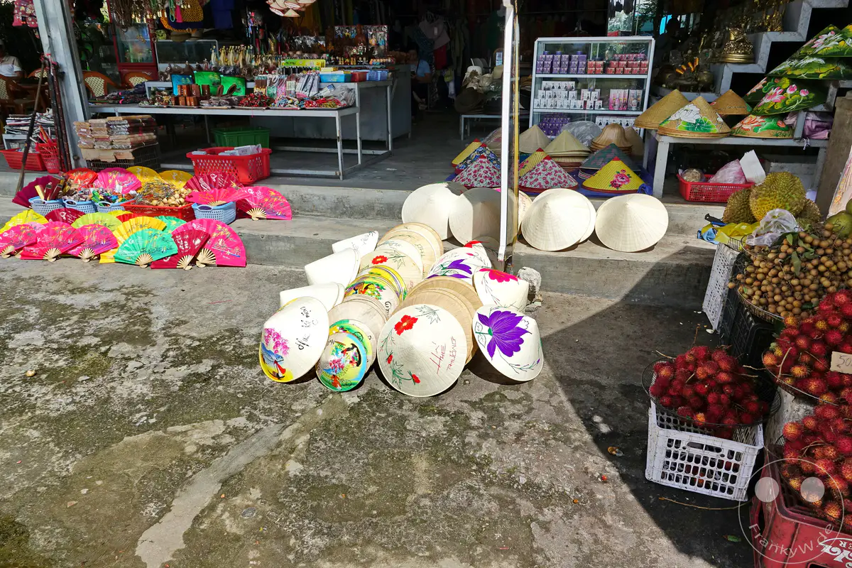 Ein lebhafter Marktstand in Hue, Vietnam, präsentiert traditionelle, handbemalte Non La Hüte und eine Vielzahl von farbenfrohen Fächern, die zum Verkauf angeboten werden. Zahlreiche Früchte wie Rambutan und Longan sind ebenfalls zu sehen und unterstreichen die Vielfalt des Angebots. Die Szenerie vermittelt einen Eindruck von der reichen Kultur und dem geschäftigen Treiben auf vietnamesischen Märkten.