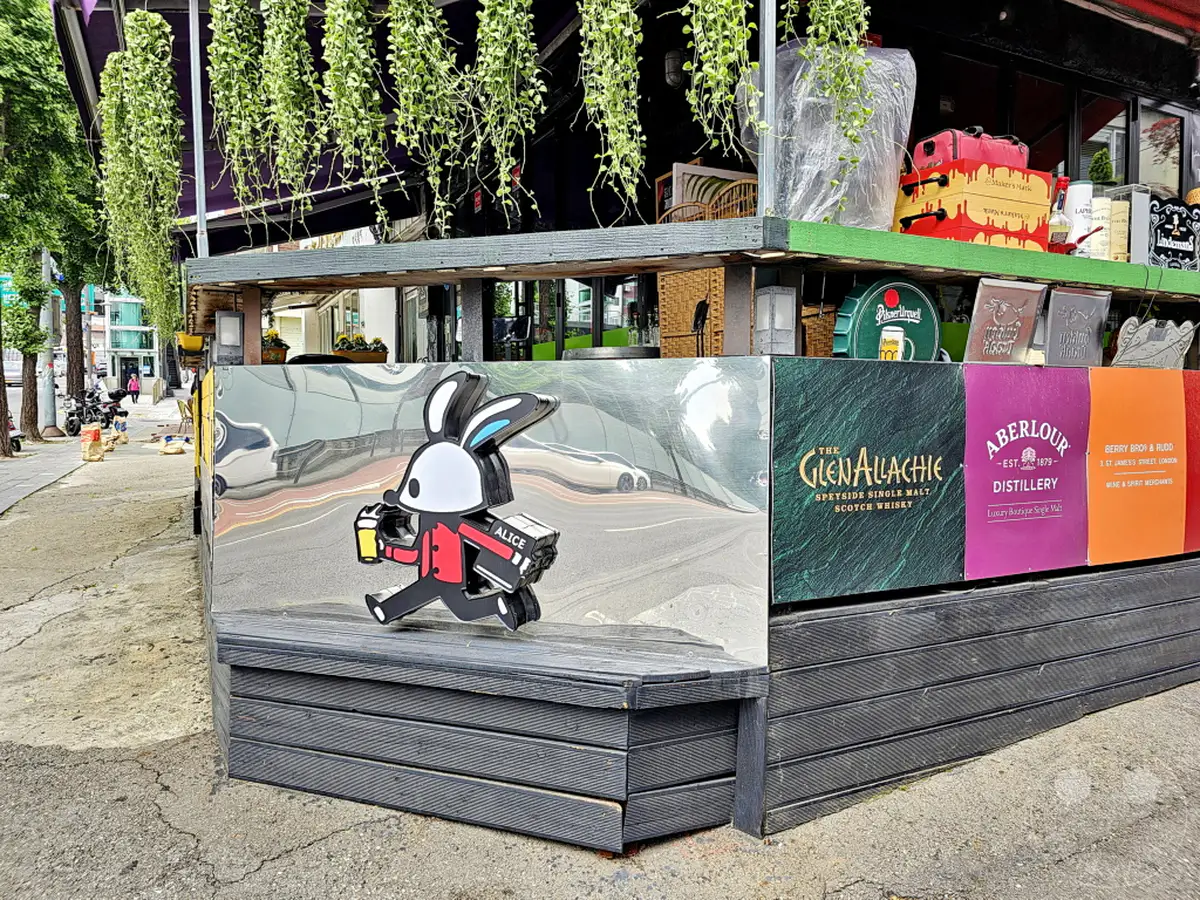 Die Außenterrasse eines Restaurants im Yongsan District in Seoul, Südkorea, präsentiert sich mit einer kreativen Dekoration. Ein beleuchteter Cartoon-Hase, der ein Getränk hält, ist auf der polierten Metallfassade angebracht, während daneben verschiedene Whisky-Flaschen und -Marken zu sehen sind. Hängende Pflanzen über dem Tresen sorgen für eine grüne und einladende Atmosphäre.