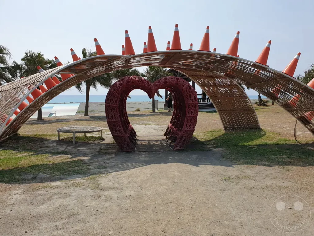 Am Dawu Coast Park in Taiwan steht eine auffällige Herzskulptur unter einer architektonischen Struktur aus Bambus und orangefarbenen Verkehrskegeln. Der Blick schweift über den Strand und das weite Meer, während Palmen im Hintergrund eine tropische Atmosphäre unterstreichen. Die Szenerie lädt zum Verweilen und Genießen der Küstenlandschaft ein.