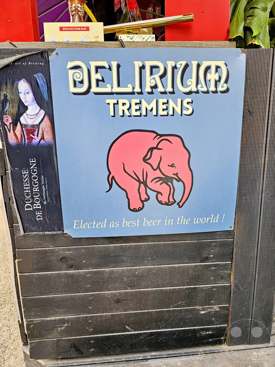 Ein Straßenschild für Delirium Tremens Bier wirbt prominent mit einem rosafarbenen Elefanten und der Aufschrift "Elected as best beer in the world!". Das Schild hängt vor einer dunkel gebeizten Holzfassade im Yongsan District von Seoul, Südkorea. Eine daneben hängende weitere Werbung zeigt eine historische Frauengestalt mit einem Vogel, was dem Ambiente eine interessante Note verleiht.