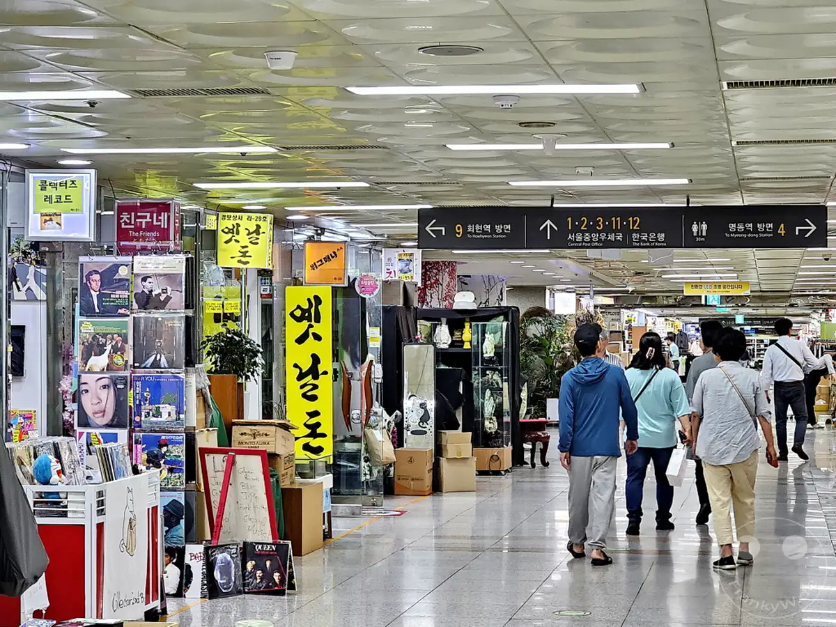 Blick in eine belebte unterirdische Einkaufspassage in der Myeongdong Metrostation in Seoul, Südkorea. An den Seiten reihen sich kleine Geschäfte aneinander, die Musik-Vinyls und andere Waren verkaufen. Menschen gehen geschäftig ihren Weg durch den Gang, belebt von Leuchtreklamen und Hinweisschildern.