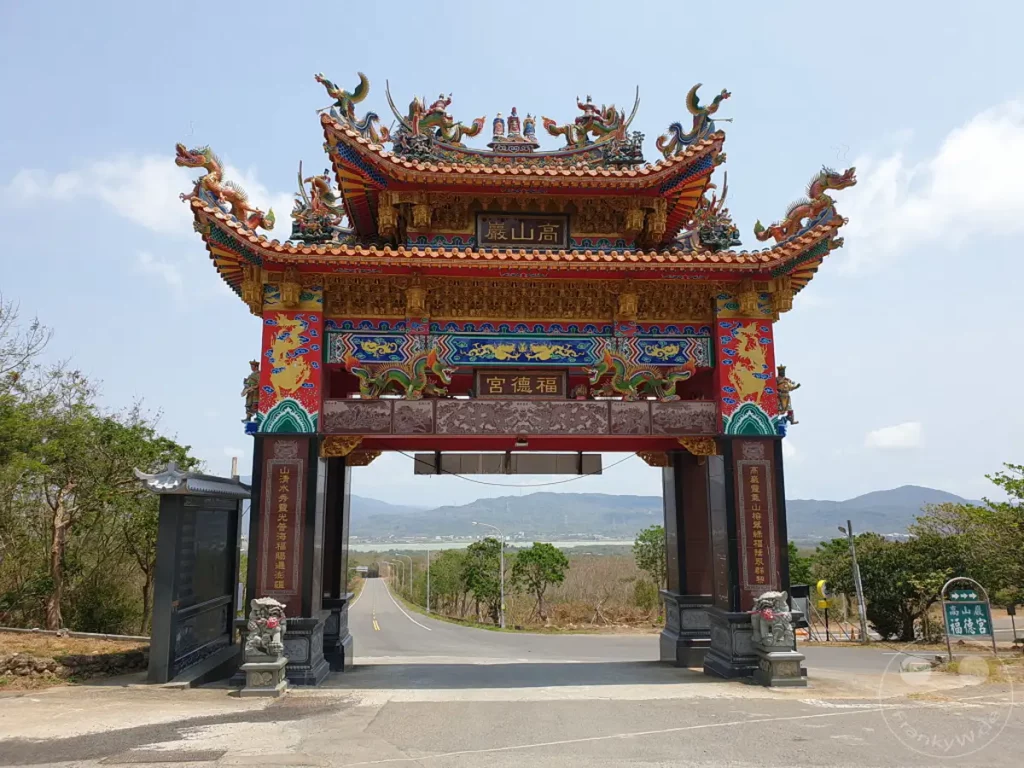 Ein prunkvolles, traditionelles taiwanisches Tempeltor steht im Vordergrund, geschmückt mit bunten Drachen und detailreicher Architektur. Die Straße führt auf dieses imposante Bauwerk zu, eingerahmt von üppiger Vegetation und einer Bergkulisse im Hintergrund. Die Szene strahlt eine Atmosphäre von kultureller Erhabenheit und friedlicher Harmonie aus.