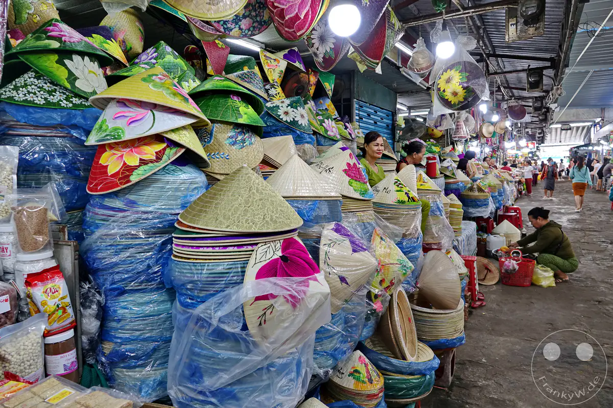 In einem geschäftigen Street Market in Hue, Vietnam, werden traditionelle, handbemalte Hüte aus Bambus und anderen Naturmaterialien zum Verkauf angeboten. Dicht aneinander gestapelte, bunte und kunstvoll verzierte Non La Hüte bilden das Herzstück des Bildes, während Menschen im Hintergrund zwischen den Ständen hindurchgehen. Die Szene strahlt die lebendige Atmosphäre eines lokalen Marktes aus, der lokale Kultur und Handwerkskunst präsentiert.