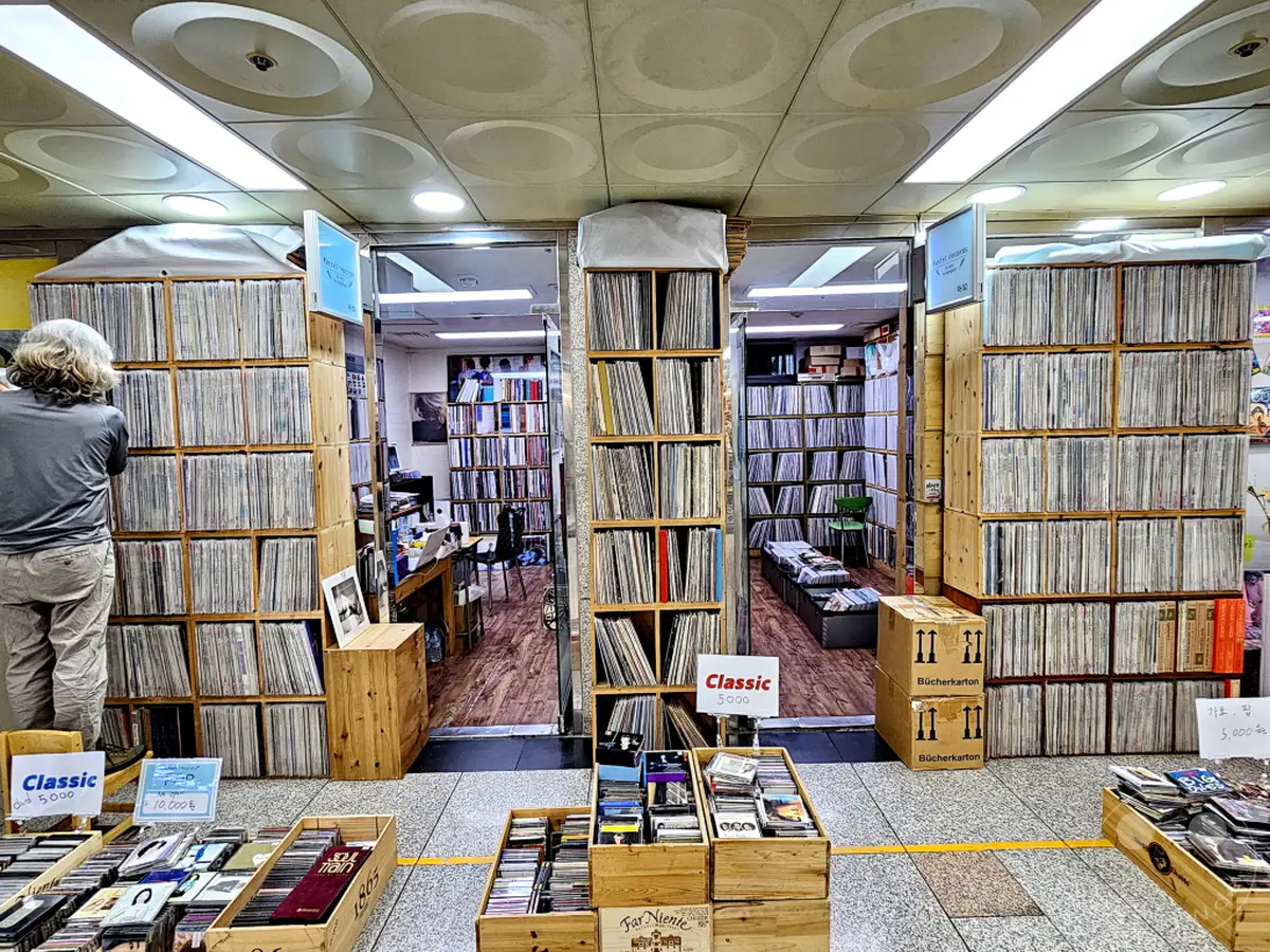 Ein Mann mit langem Haar steht in einem vollgestopften Geschäft, das an einer U-Bahn-Station liegt und Schallplatten und CDs verkauft. Regale voller Vinyl-Schallplatten säumen die Wände und bilden schmale Gänge, während Holzkisten mit CDs im Vordergrund auf dem Boden stehen. Die Atmosphäre ist geschäftig und lädt dazu ein, in der Musiksammlung zu stöbern.