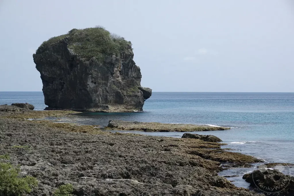 Der markante Sail Rock ragt imposant aus dem Meer vor der Küste Taiwans auf. Die felsige Uferlinie im Vordergrund bildet einen rauen Kontrast zum ruhigen, türkisblauen Meer und dem hellen Himmel. Die Szenerie vermittelt eine friedliche und natürliche Atmosphäre, die zum Verweilen einlädt.