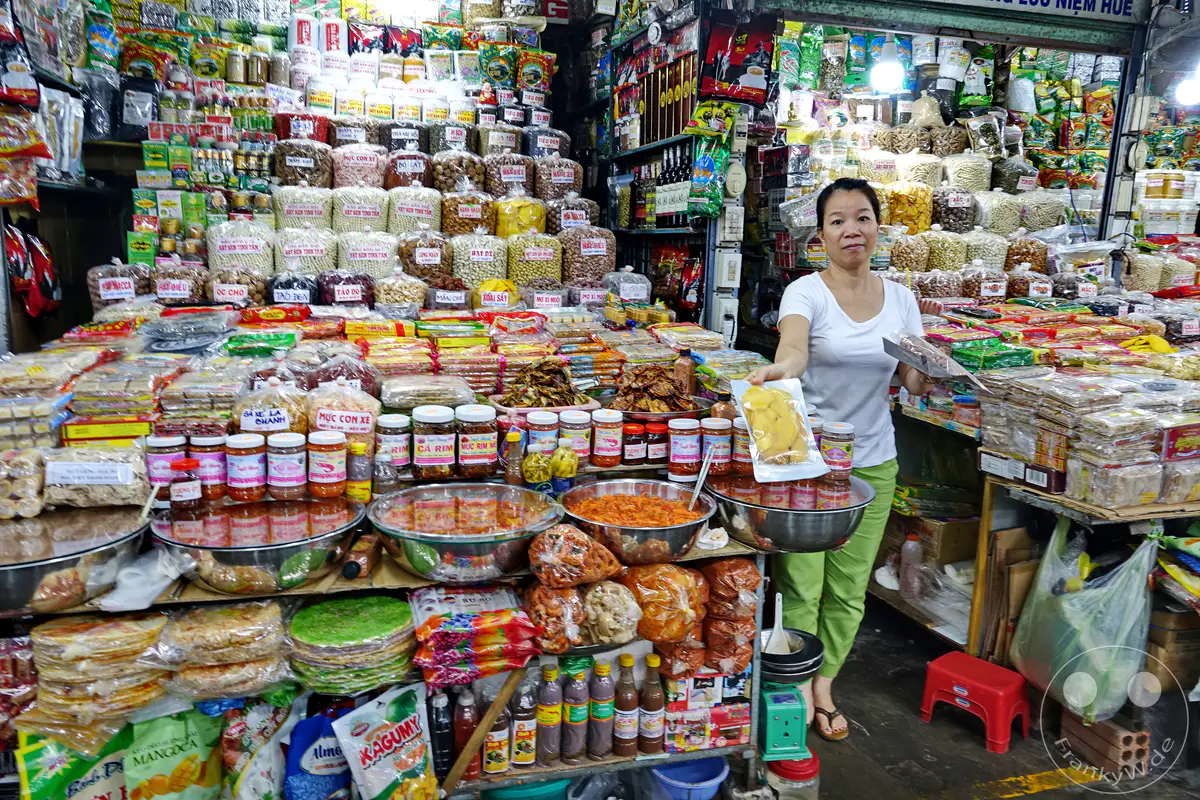 Ein lebhafter Street Market in Hue, Vietnam, präsentiert eine Fülle von Lebensmitteln und Gewürzen, die in bunten Verpackungen und Gläsern gestapelt sind. Eine Verkäuferin bietet mehrere Produkte an, während die Waren eine reiche Vielfalt an kulinarischen Traditionen widerspiegeln. Die Szene strahlt die geschäftige Energie und den authentischen Charme eines lokalen Marktes aus.