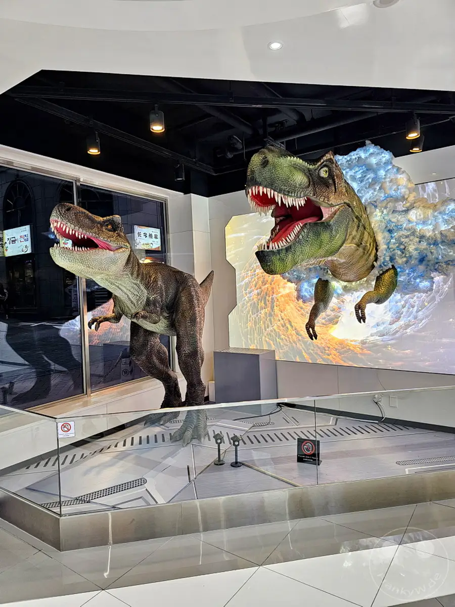 Zwei lebensgroße Dinosaurier-Nachbildungen, eine davon durchbricht eindrucksvoll eine beleuchtete Wand, sind das Herzstück einer faszinierenden Ausstellung. Die Szenerie spielt sich im Eingangsbereich des Henn-na Hotels im belebten Myeongdong, Seoul, ab und verspricht ein prähistorisches Abenteuer für Besucher. Ein klares Glaselement trennt die Besucher von den Exponaten und unterstreicht den musealen Charakter der Darbietung.