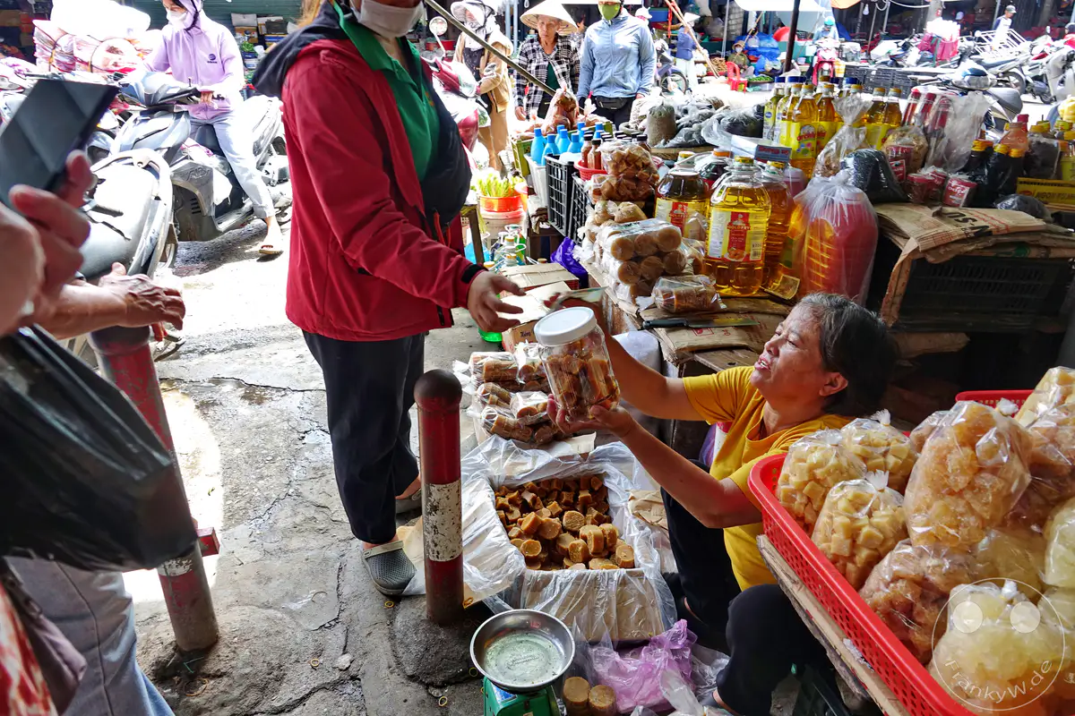 Ein geschäftiger Marktstand in Hue, Vietnam, bietet eine Vielzahl von Lebensmitteln wie Zuckerrohrzucker und Gewürze an. Händlerinnen und Kunden interagieren inmitten des bunten Treibens, während Motorroller und Verkaufsstände das Bild prägen. Die Szene vermittelt die lebendige Atmosphäre eines traditionellen Marktes im südostasiatischen Raum.