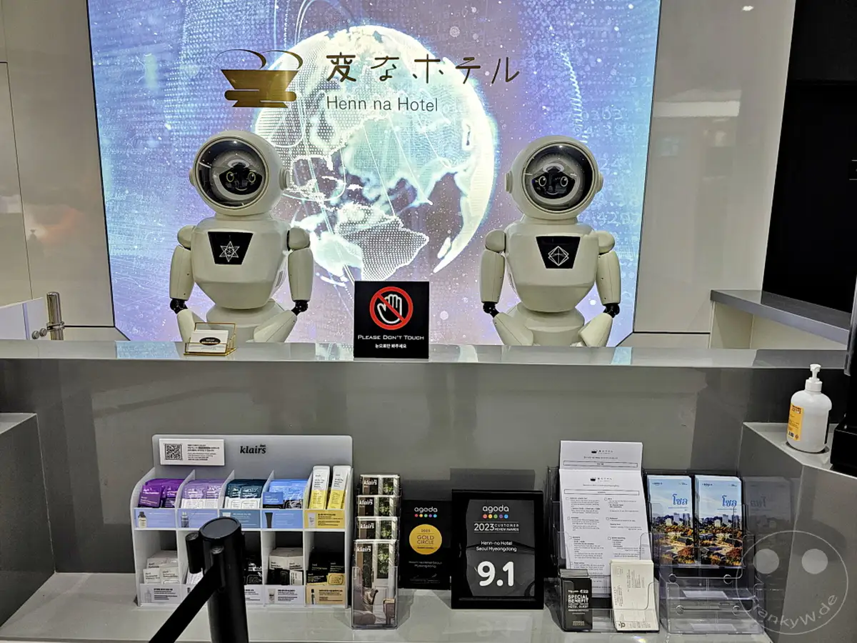 Zwei humanoide Roboter posieren hinter einer Rezeption in Südkorea, genauer gesagt im Henn na Hotel in Seoul, Myeongdong. Auf dem Tresen sind verschiedene Informationsmaterialien und Produkte ausgestellt, während im Hintergrund eine Weltkugel-Grafik zu sehen ist. Die Szene vermittelt eine futuristische und technologisch fortschrittliche Atmosphäre.