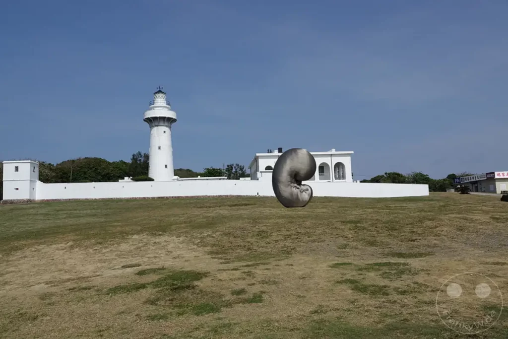 Am Eluanbi Lighthouse auf Taiwan steht eine markante, graue Skulptur vor dem weißen historischen Leuchtturm und einer niedrigen Mauer. Die Szene wird von einer weiten, trockenen Grasfläche dominiert, die sich bis zum Horizont erstreckt, unter einem klaren, blauen Himmel. Die weite und offene Landschaft strahlt eine ruhige und sonnige Atmosphäre aus, die die Architektur und die künstlerische Installation hervorhebt.
