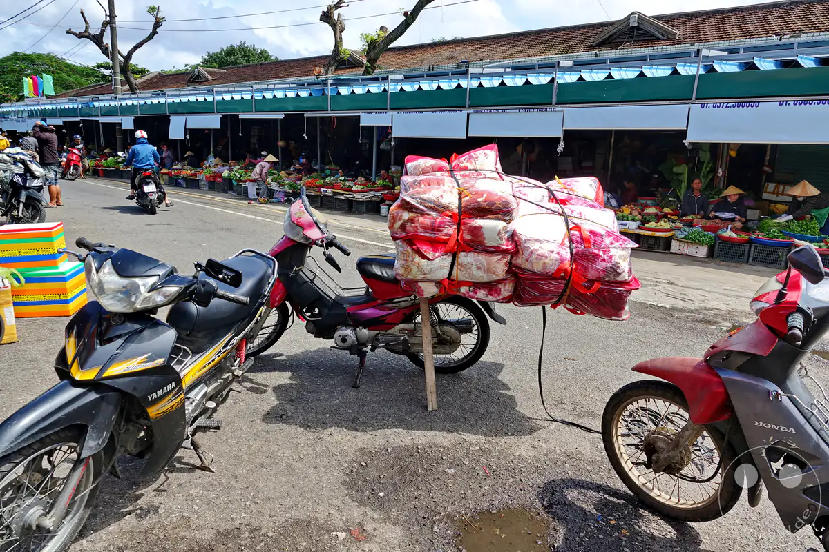 Auf einem belebten Markt in Hue, Vietnam, stehen mehrere Motorroller bereit, um Waren zu transportieren. Ein Roller ist mit einer großen Menge an Decken beladen, die für den Verkauf oder die Lieferung bestimmt sind. Die Szene zeigt das geschäftige Treiben eines lokalen Marktes, wo Handel und Verkehr auf den Straßen eng miteinander verbunden sind.