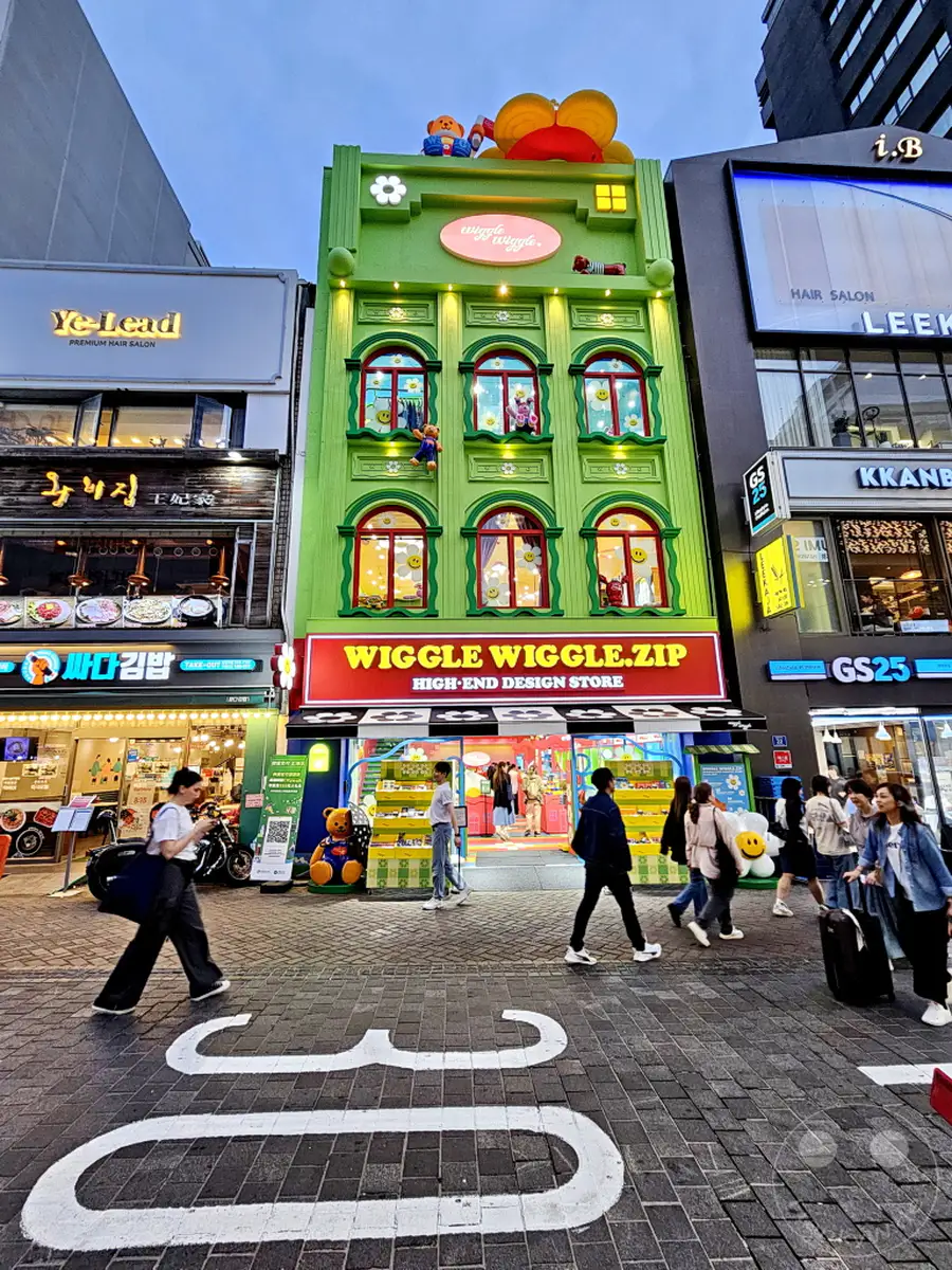 Eine farbenfrohe Fassade des "Wiggle Wiggle Store" dominiert das Bild in Südkorea, Seoul. Menschen spazieren auf einer belebten Straße, die mit auffälligen weißen Bodenmarkierungen versehen ist. Die Szene strahlt eine lebendige und verspielte Atmosphäre aus, die typisch für die Einkaufsgegend von Myeongdong ist.