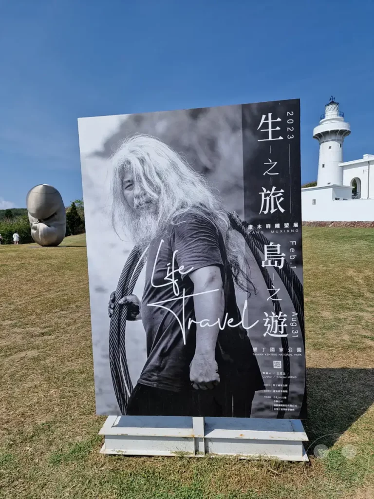 Eine große Ausstellungsplakat mit einem Porträt des Künstlers Kang Muxiang steht im Vordergrund und zeigt die Ausstellung "Leben - Reise". Im Hintergrund ist der ikonische Eluanbi Lighthouse auf Taiwan zu sehen, der über einer grasbewachsenen Hügelkuppe thront. Die Szene vermittelt eine ruhige und künstlerische Atmosphäre unter blauem Himmel.