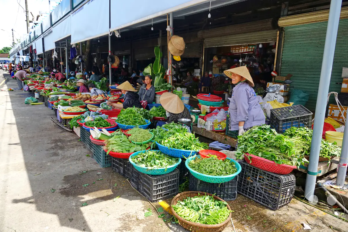Ein lebhafter Street Market in Hue, Vietnam, präsentiert eine Fülle von frischem Gemüse, das von Händlerinnen in traditionellen asiatischen Hüten angeboten wird. Die Szene strahlt die geschäftige Atmosphäre eines lokalen Marktes aus, wo bunte Waren auf dem Gehweg ausgelegt sind. Es ist ein authentischer Einblick in das tägliche Leben und die kulinarische Vielfalt der Stadt.