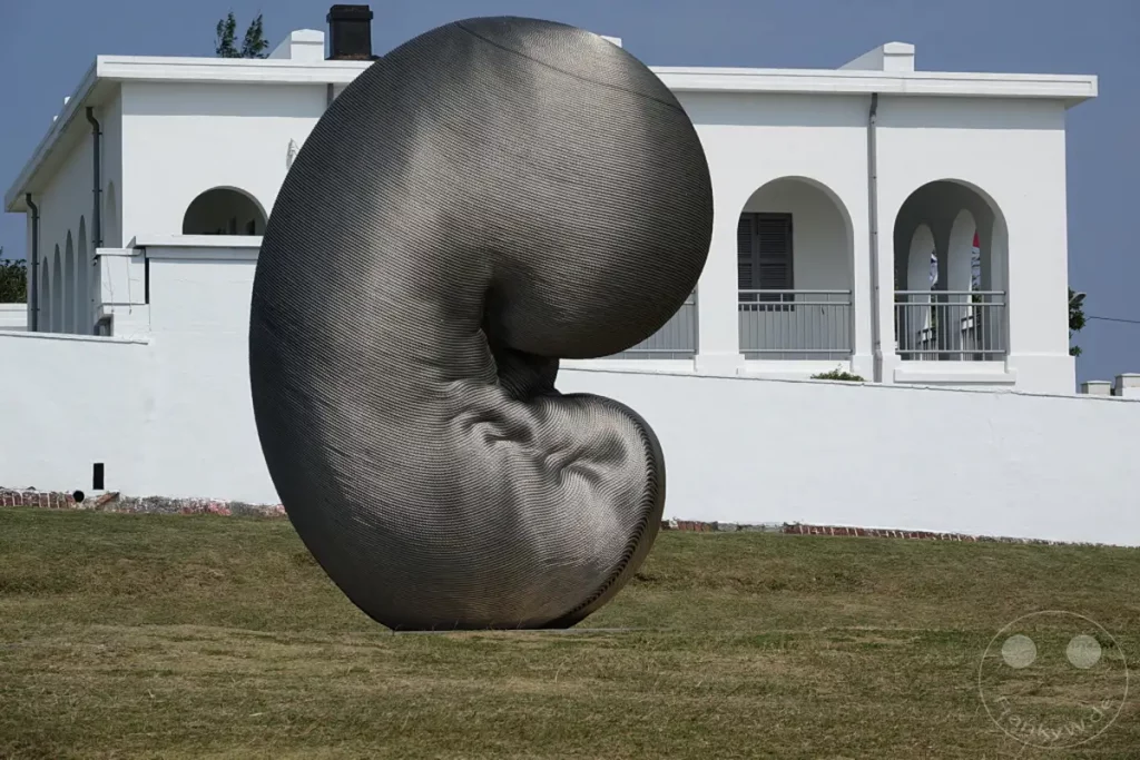Vor dem markanten weißen Gebäude des Eluanbi Leuchtturms in Taiwan erhebt sich die beeindruckende Skulptur "Rebirth" von Kang Muxiang. Die organisch geformte Metallkonstruktion steht auf einer gepflegten Rasenfläche und bildet einen faszinierenden Kontrast zur klaren Architektur im Hintergrund. Die strahlende Sonne und der blaue Himmel verleihen der Szene eine ruhige und imposante Atmosphäre.
