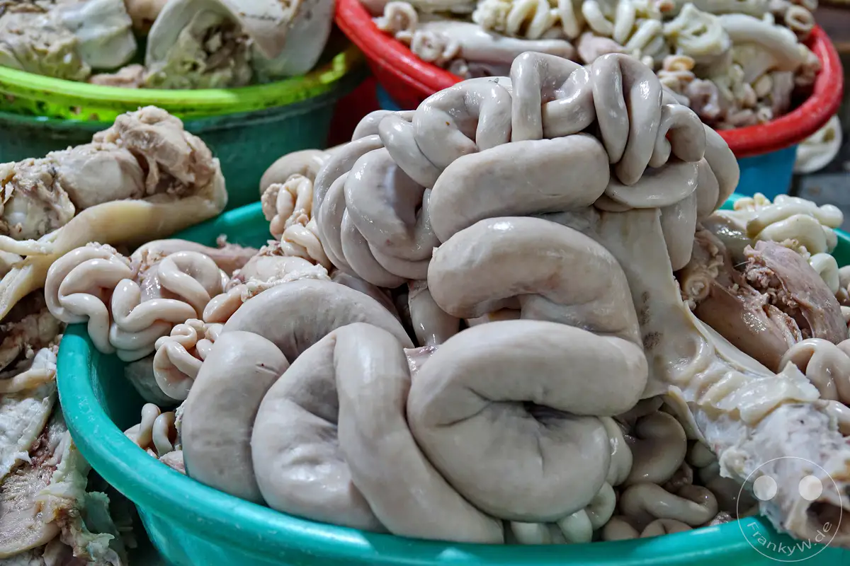 In einem Markt in Hue, Vietnam, werden frisch zubereitete Schweineinnereien in einem tiefen, türkisfarbenen Plastikbehälter präsentiert. Die gekochten oder blanchierten Innereien sind in verschiedenen Formen und Größen zu sehen und werden von weiteren Portionen in anderen Behältern im Hintergrund ergänzt. Die Nahaufnahme lässt die Textur und Frische der Ware erkennen, die auf einem typischen Street Market angeboten wird.