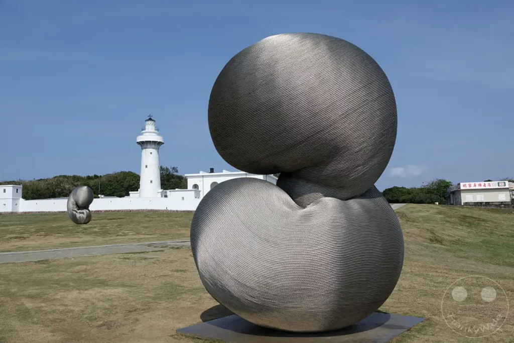 Taiwan - Eluanbi Lighthouse - Kang Muxiang - Sculpture Rebirth - Art Installation Die markante Skulptur "Rebirth" von Kang Muxiang steht vor dem Eluanbi Lighthouse in Taiwan. Die Installation besteht aus zwei metallischen, abstrakten Formen, die sich organisch aneinanderfügen. Im Hintergrund erhebt sich das weiße, zylindrische Bauwerk des Leuchtturms unter einem strahlend blauen Himmel, umgeben von grünen Hügeln und dem Meer.
