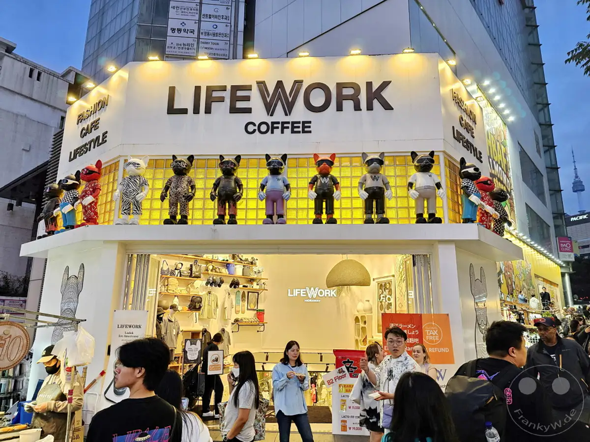 Die "LIFEWORK COFFEE" Filiale in Myeongdong, Seoul, präsentiert sich mit bunten Figuren auf dem Vordach und einem stilvollen Eingangsbereich. Die Fassade des Modegeschäfts und Cafés zieht mit seinem auffälligen Design und den Dekorationen die Aufmerksamkeit der Passanten auf sich. Eine belebte Atmosphäre herrscht auf der Straße vor dem Geschäft, wo Menschen einkaufen und die Stadt erkunden.