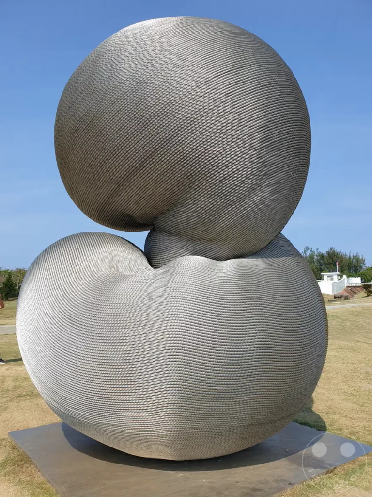 Eine monumentale Skulptur aus Aluminiumseilen, "Rebirth" von Kang Muxiang, steht im Freien nahe dem Eluanbi Leuchtturm in Taiwan. Die organischen Formen der Kunstinstallation ähneln sich überlagernden Blasen oder tropfenförmigen Elementen, die im Sonnenlicht glänzen. Die weite Grünfläche und der klare blaue Himmel schaffen eine ruhige und erhabene Atmosphäre für dieses beeindruckende Kunstwerk.
