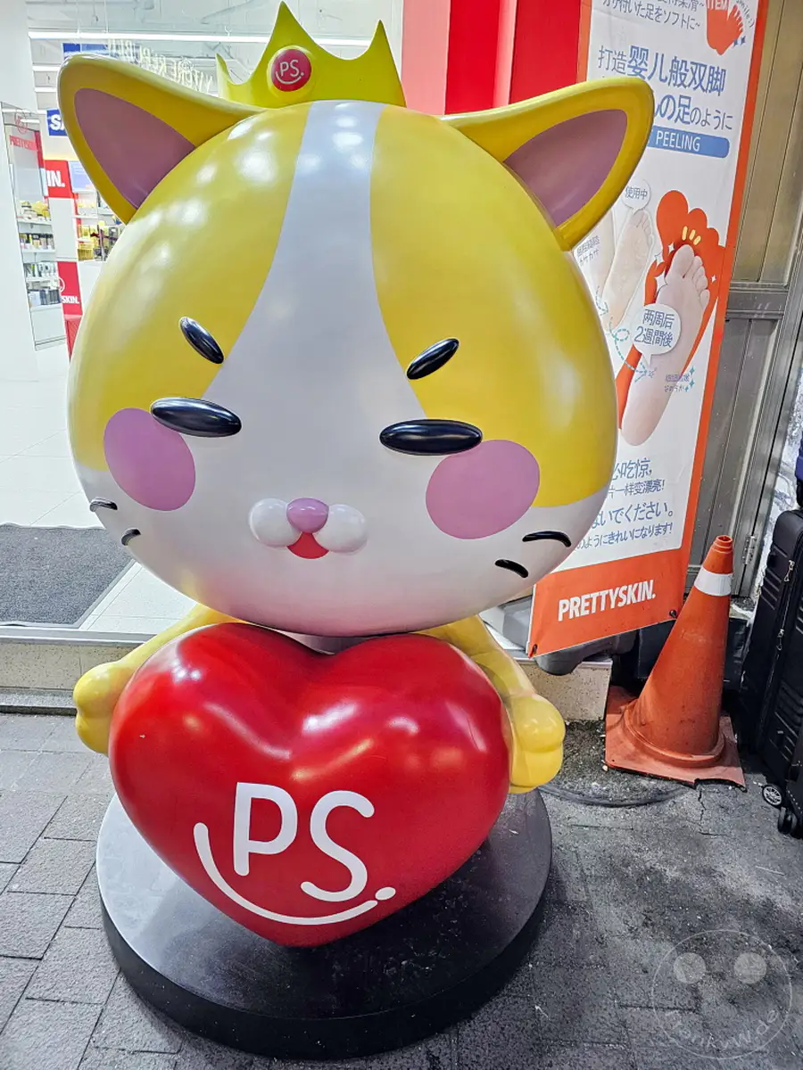 Eine lebensgroße, gelb-weiße Katzenfigur mit Krone und einem roten Herz auf dem Bauch steht auf dem Bürgersteig in Südkorea. Sie wirbt für die Kosmetikmarke 'PrettySkin' auf einem Plakat im Hintergrund. Die Szene vermittelt eine fröhliche und bunte Atmosphäre in der belebten Einkaufsstraße von Myeongdong, Seoul.